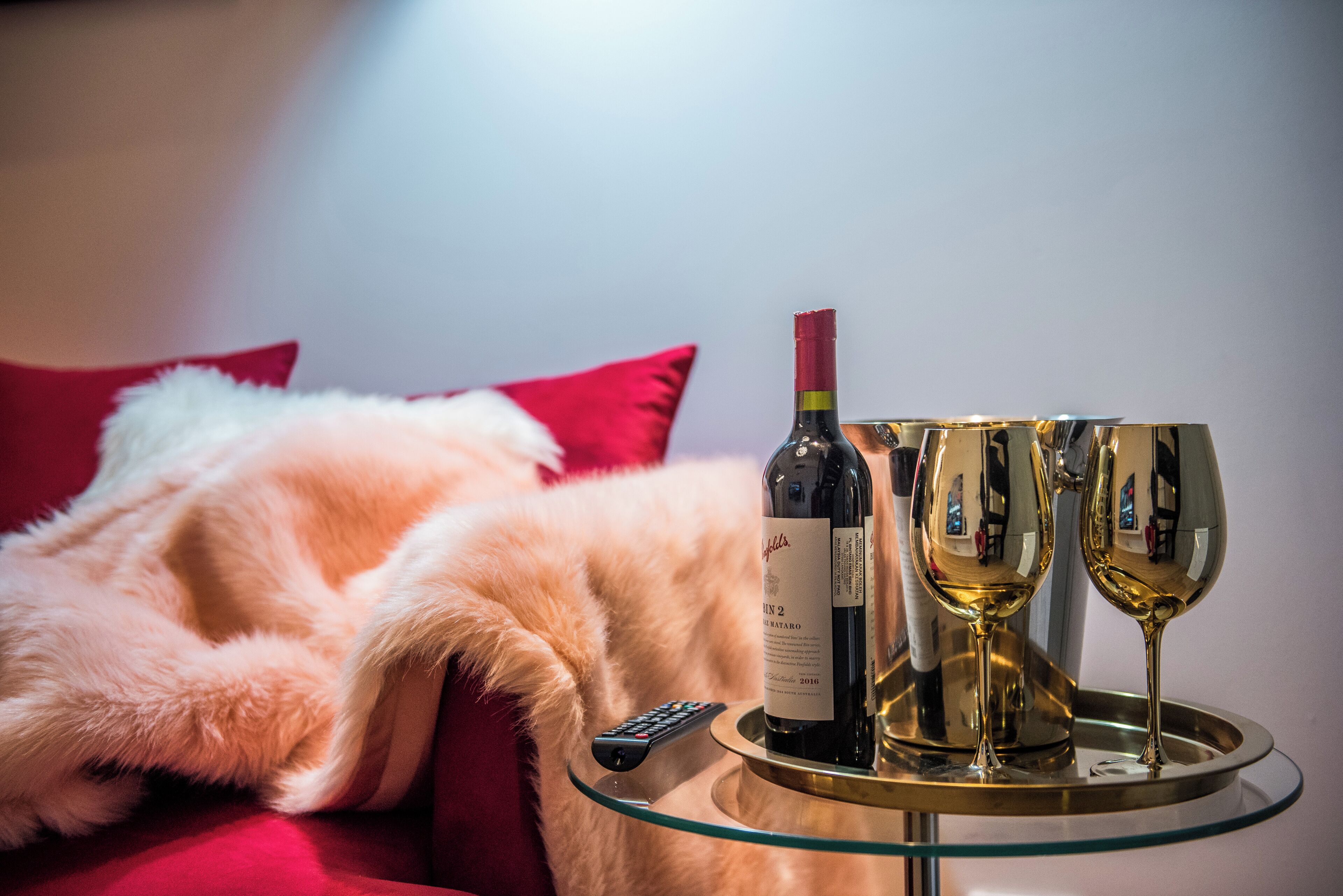 Een bed met een bontdeken, een afstandsbediening, een fles wijn en twee glaasjes op een glazen tafel.