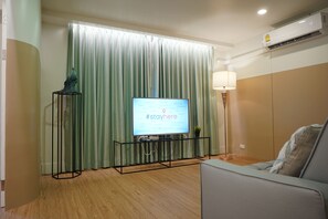 A modern living room with a flat-screen TV on a glass table, a green curtain, a lamp, and a sofa.