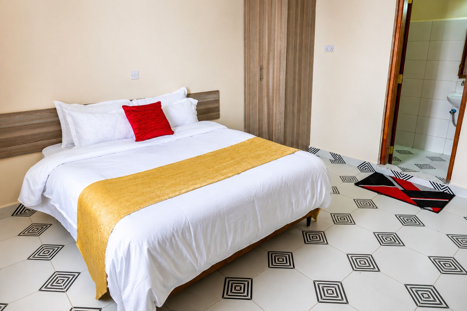 Une chambre d’hôtel avec un lit, un oreiller rouge, un plaid jaune et des carreaux de sol à motifs.