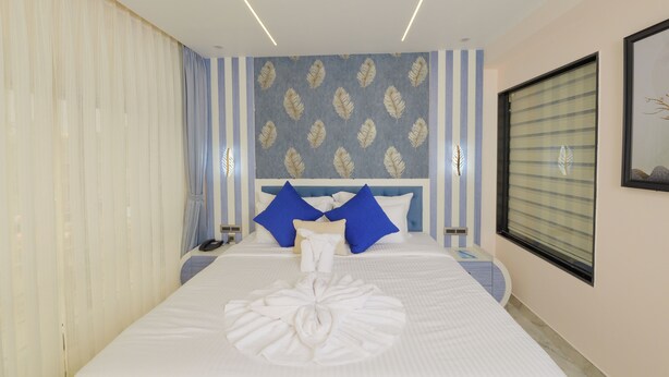 Una habitación de hotel con una cama bien hecha, un cabecero con paneles decorativos, una mesita de noche con una lámpara y una ventana con persianas.