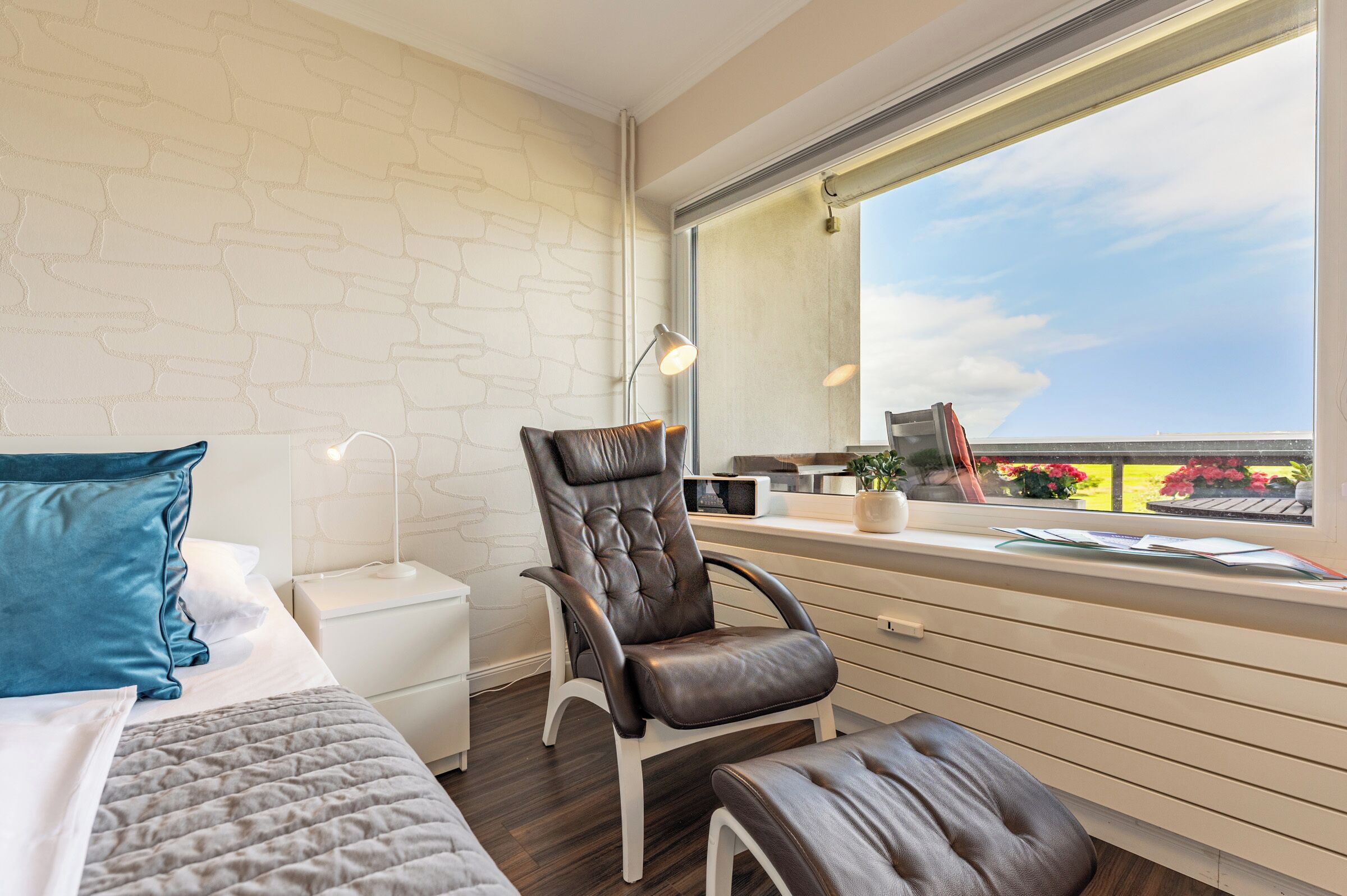 Une chambre à coucher moderne, avec un lit, une chaise, une table de chevet, une lampe et une fenêtre donnant sur un beau paysage.