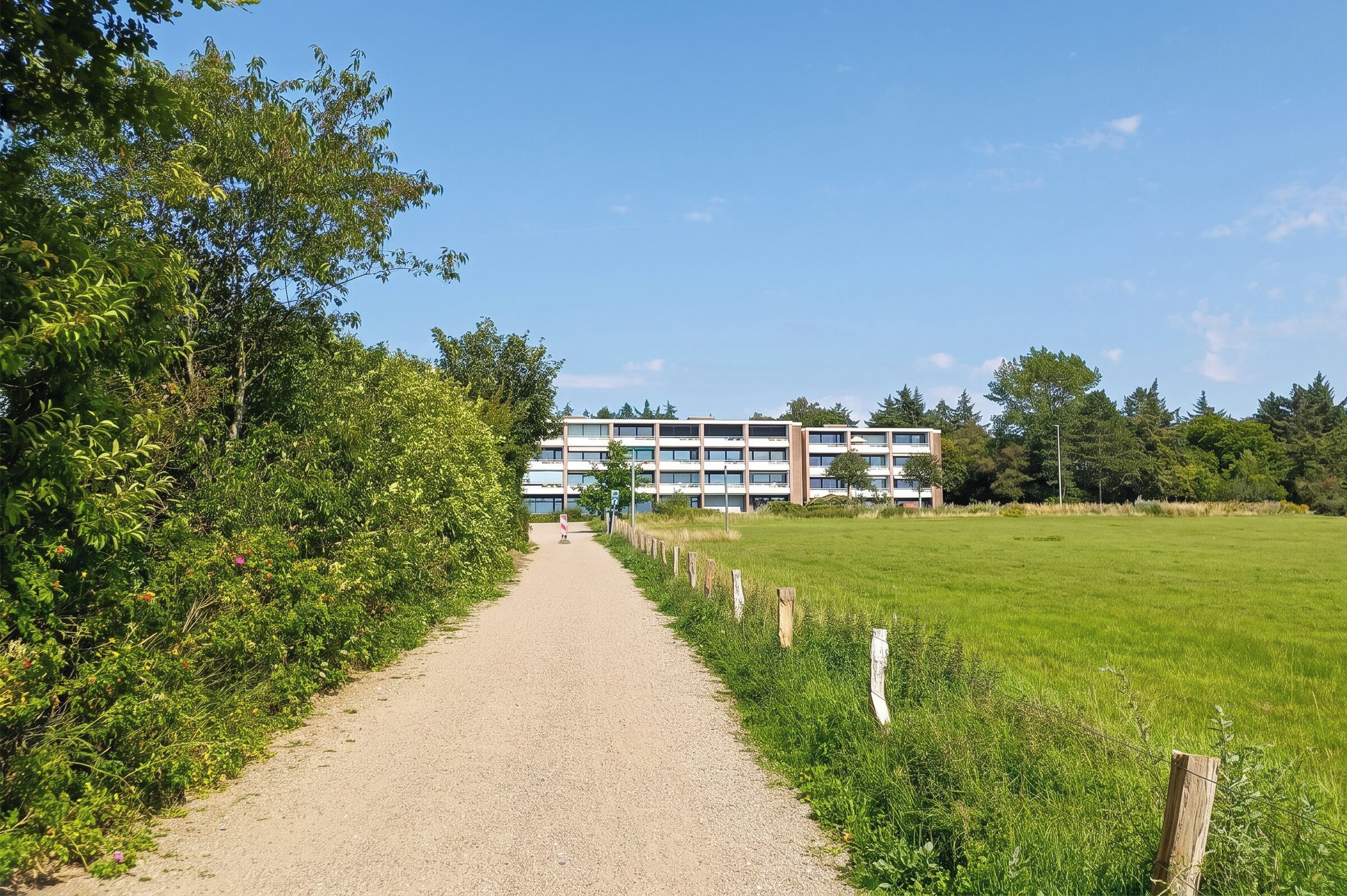 Ein Kiesweg führt zu einem modernen Gebäude mit mehreren Stockwerken und großen Fenstern.