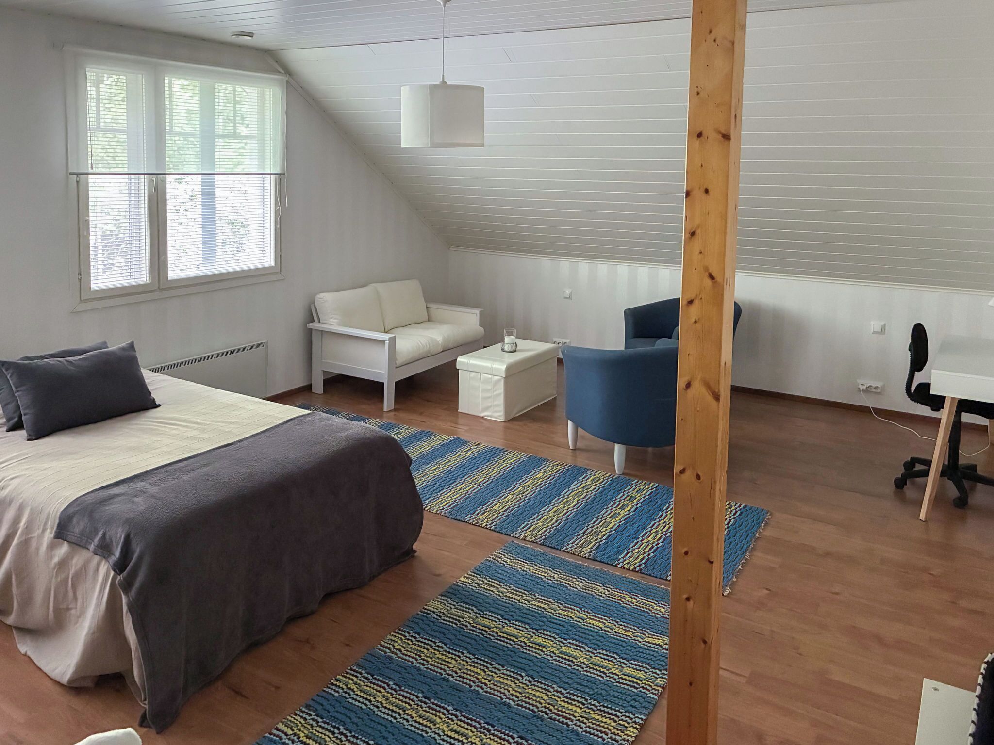 A bedroom with a bed, a white sofa, a blue chair, a desk, and a wooden beam structure.