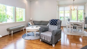 A spacious living room with a grey sofa, a round coffee table, and a bench. There are large windows providing natural light and a view of greenery outside.