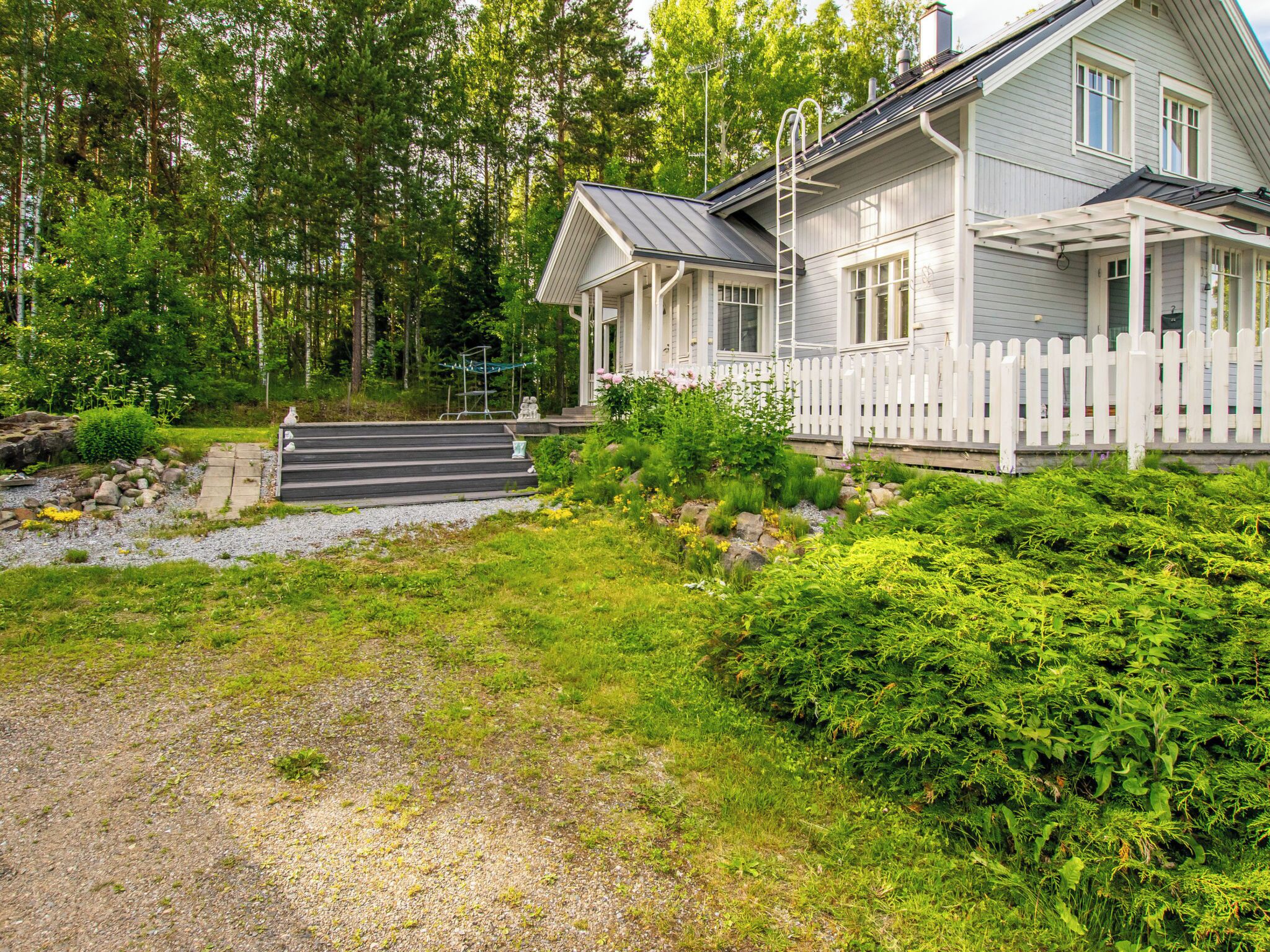 A two-story house with a porch, a gravel pathway, and a forested area.
