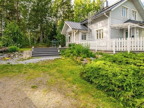 A two-story house with a porch, a gravel pathway, and a forested area.