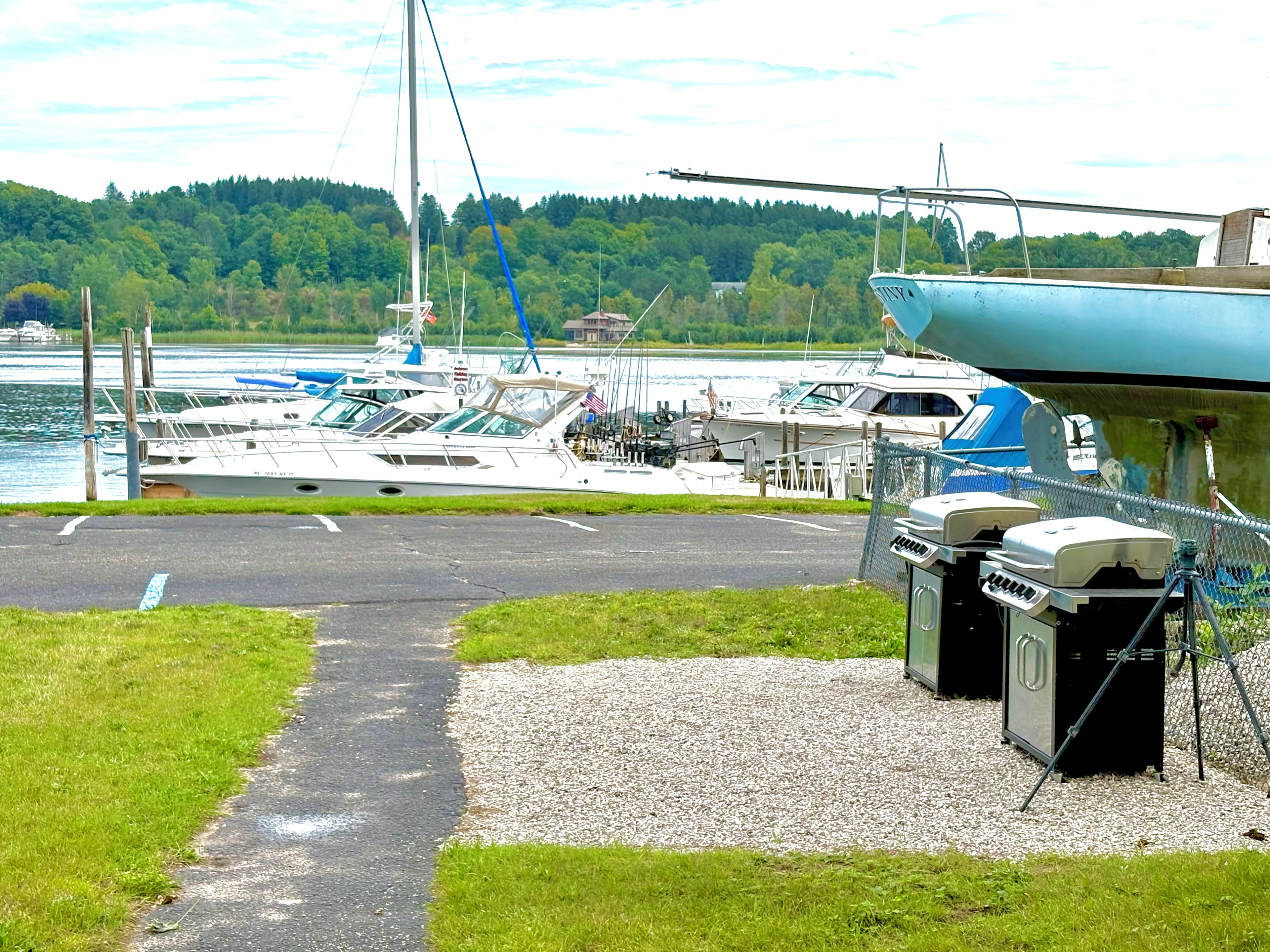 A marina with boats docked, a grassy area, and a gravel path leading to a covered area with barbecue grills.