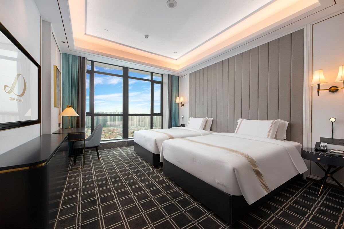 A hotel room with two beds, a desk, a chair, a large window with a city view, and a wall-mounted lamp.