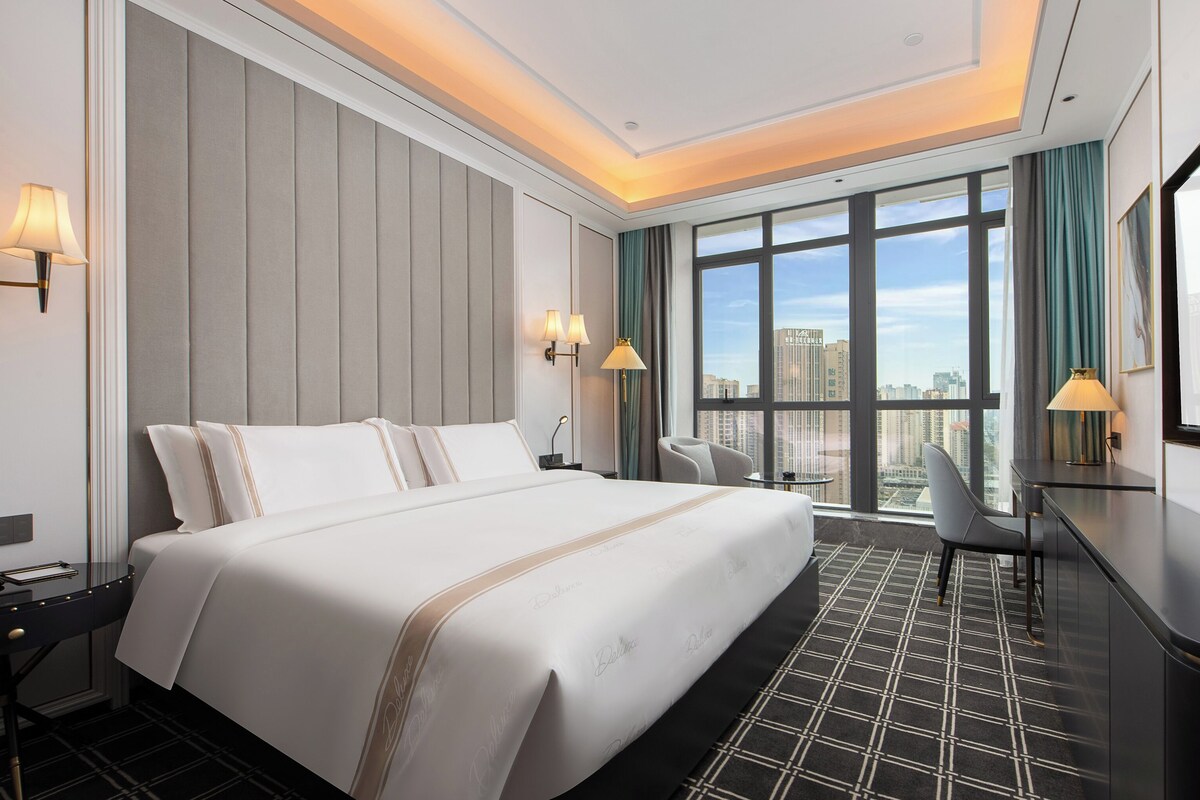 A hotel room with a large bed, a desk, and a view of the city through large windows.