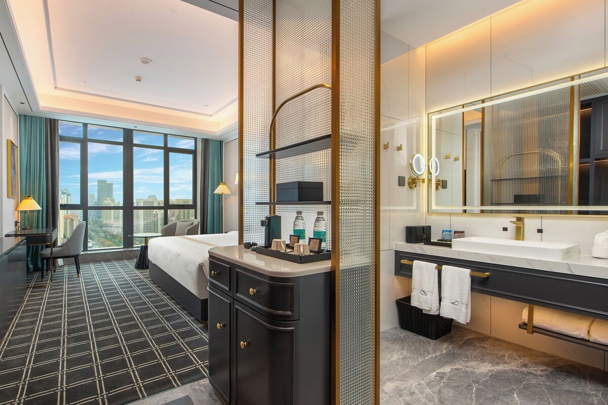 A hotel room with a large bed, a desk, a chair, a bathroom with a mirror and sink, and a city view through large windows.