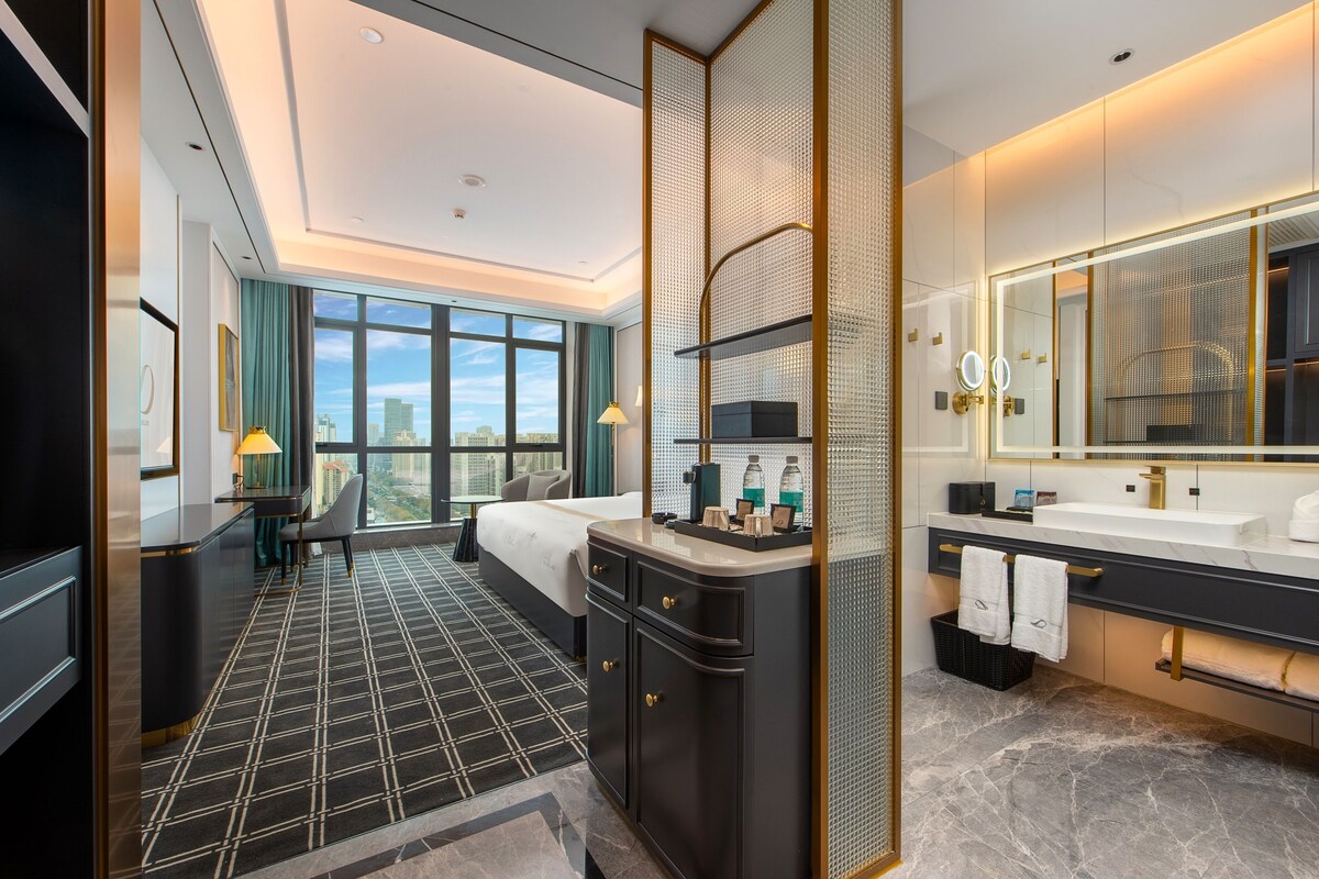 A modern hotel room with a large bed, a separate bathroom, a city view, and a check-in desk.