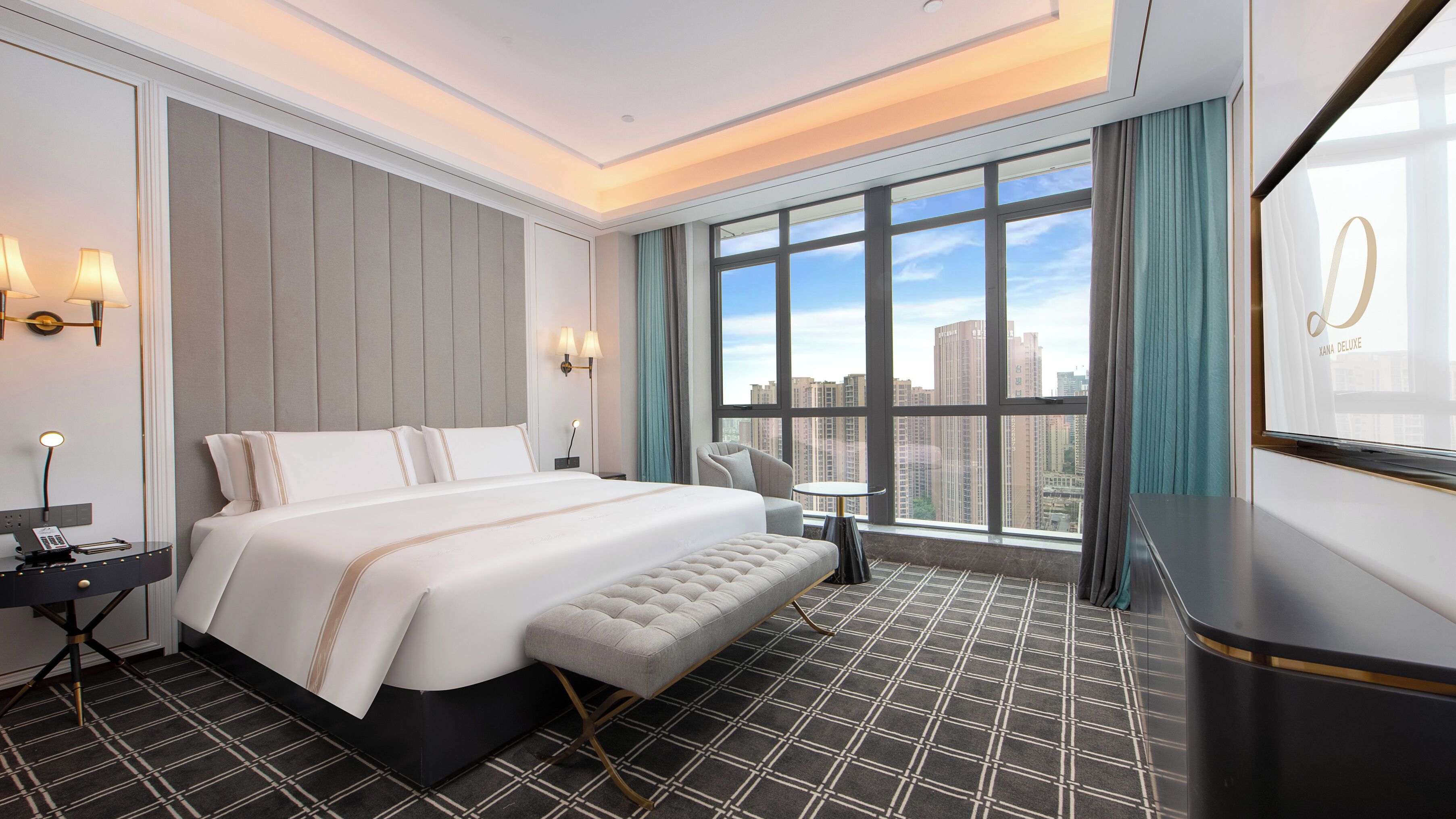 Una habitación de hotel moderna con una cama grande, zona de estar, una mesita y vistas a la ciudad a través de amplios ventanales.