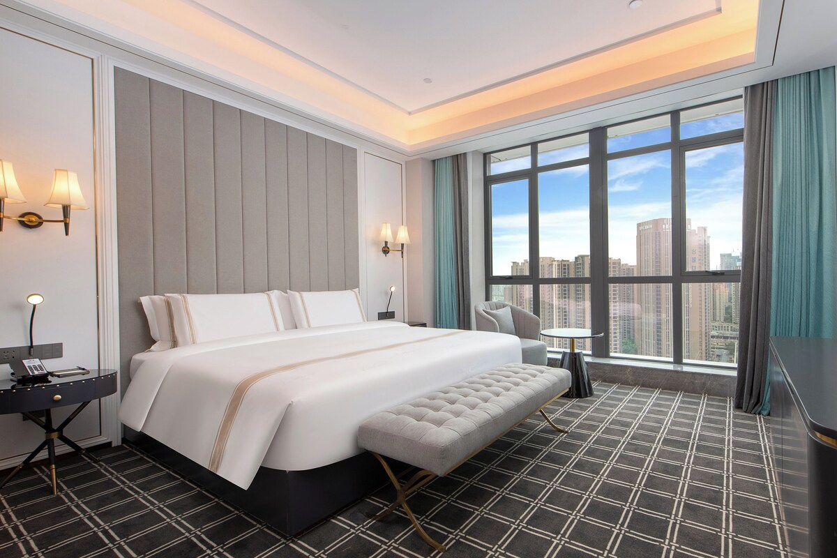 A modern hotel room with a large bed, a seating area, a small table, and a view of the cityscape through large windows.