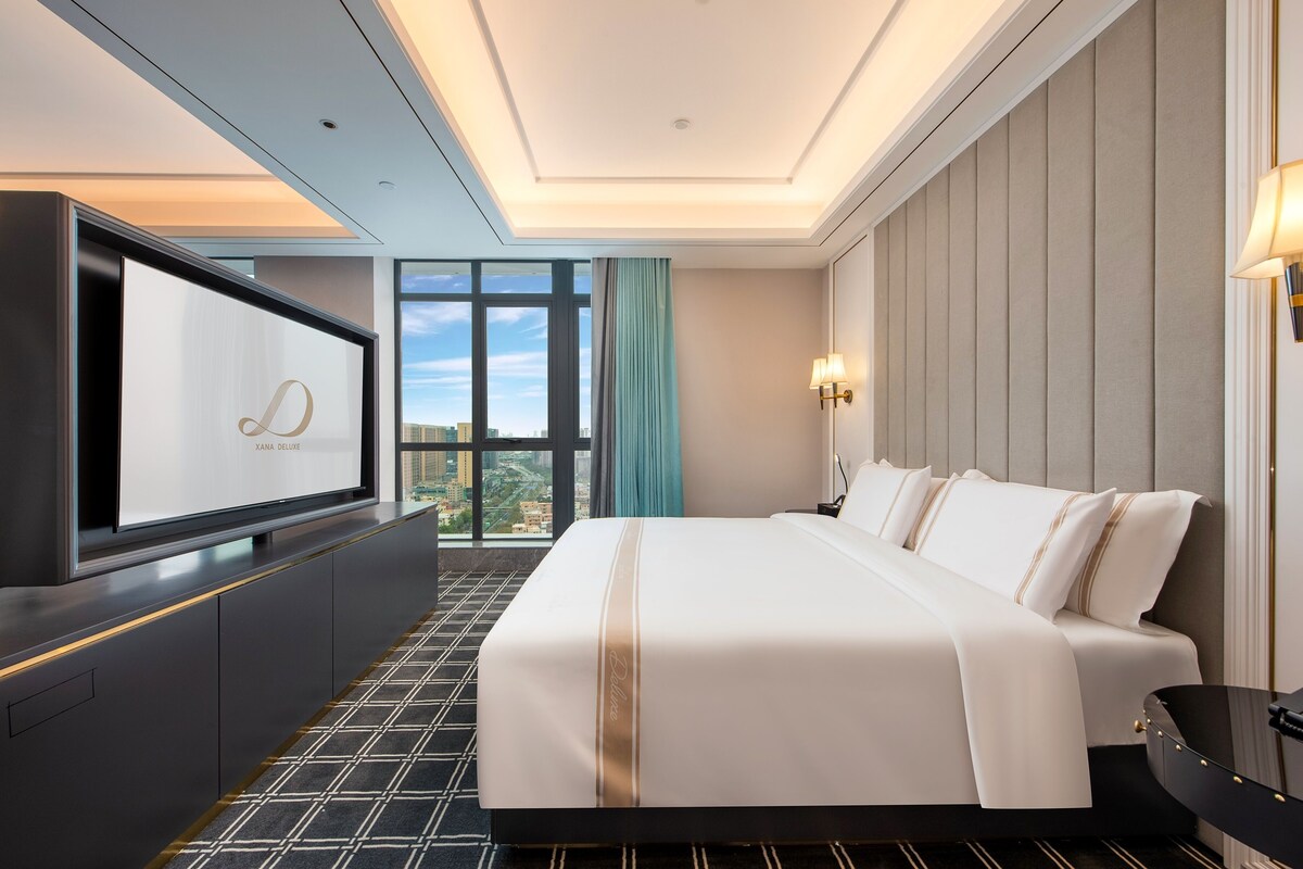 A hotel room with a large bed, a television, a view of the city, and a window with curtains.