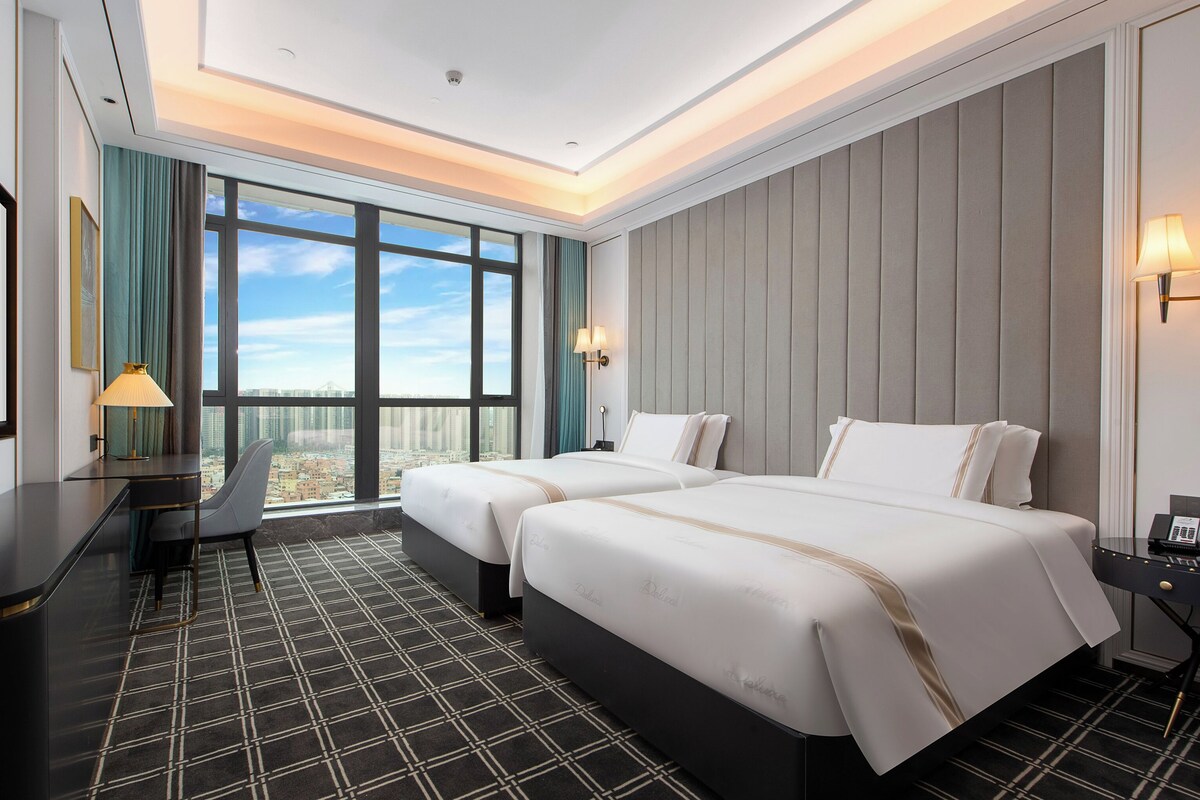 A hotel room with two beds, a desk, a chair, a large window with a city view, and a patterned floor.