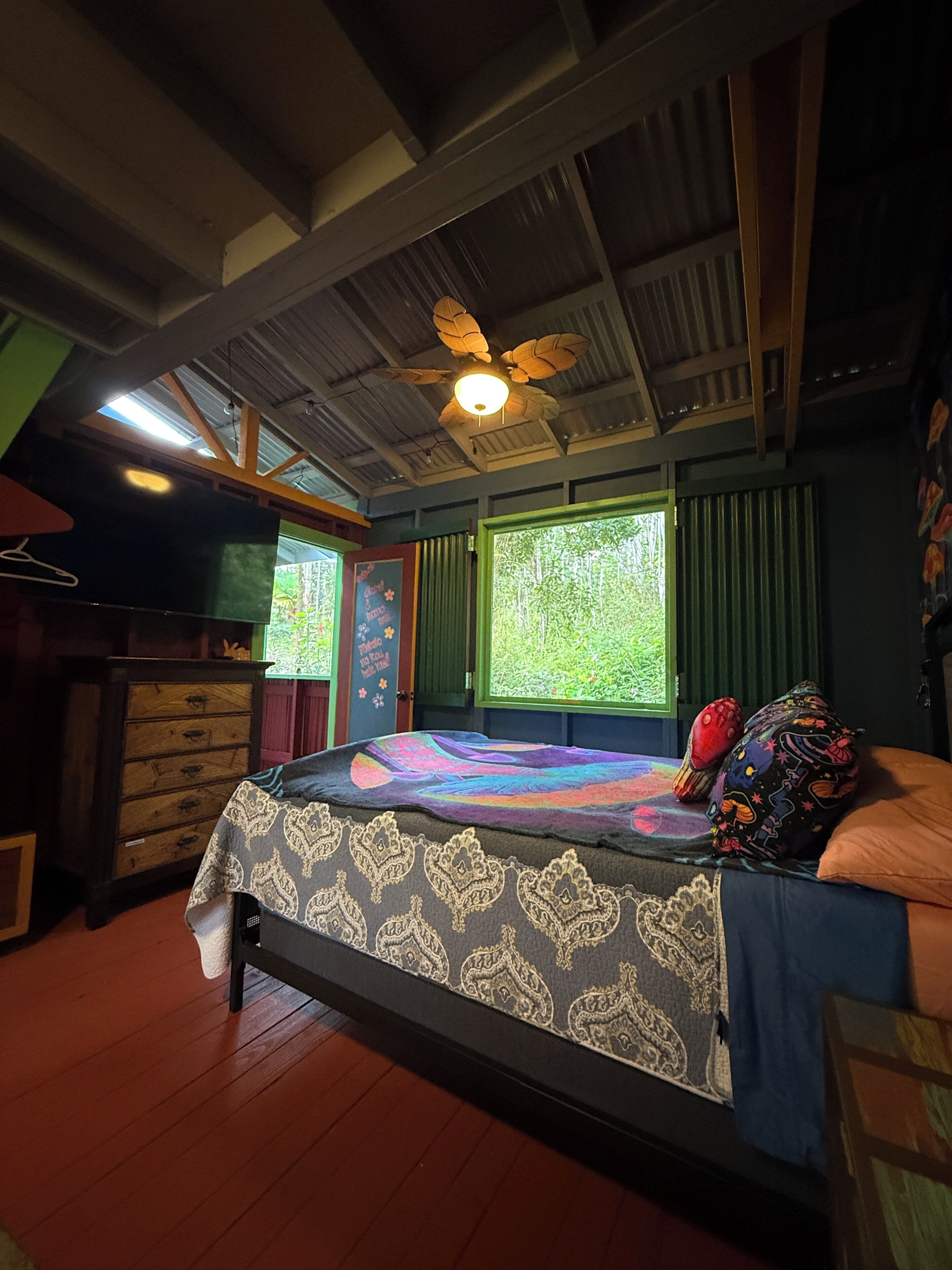 Une chambre à coucher comprenant un lit, un ventilateur de plafond, une commode et une fenêtre donnant sur un paysage verdoyant.