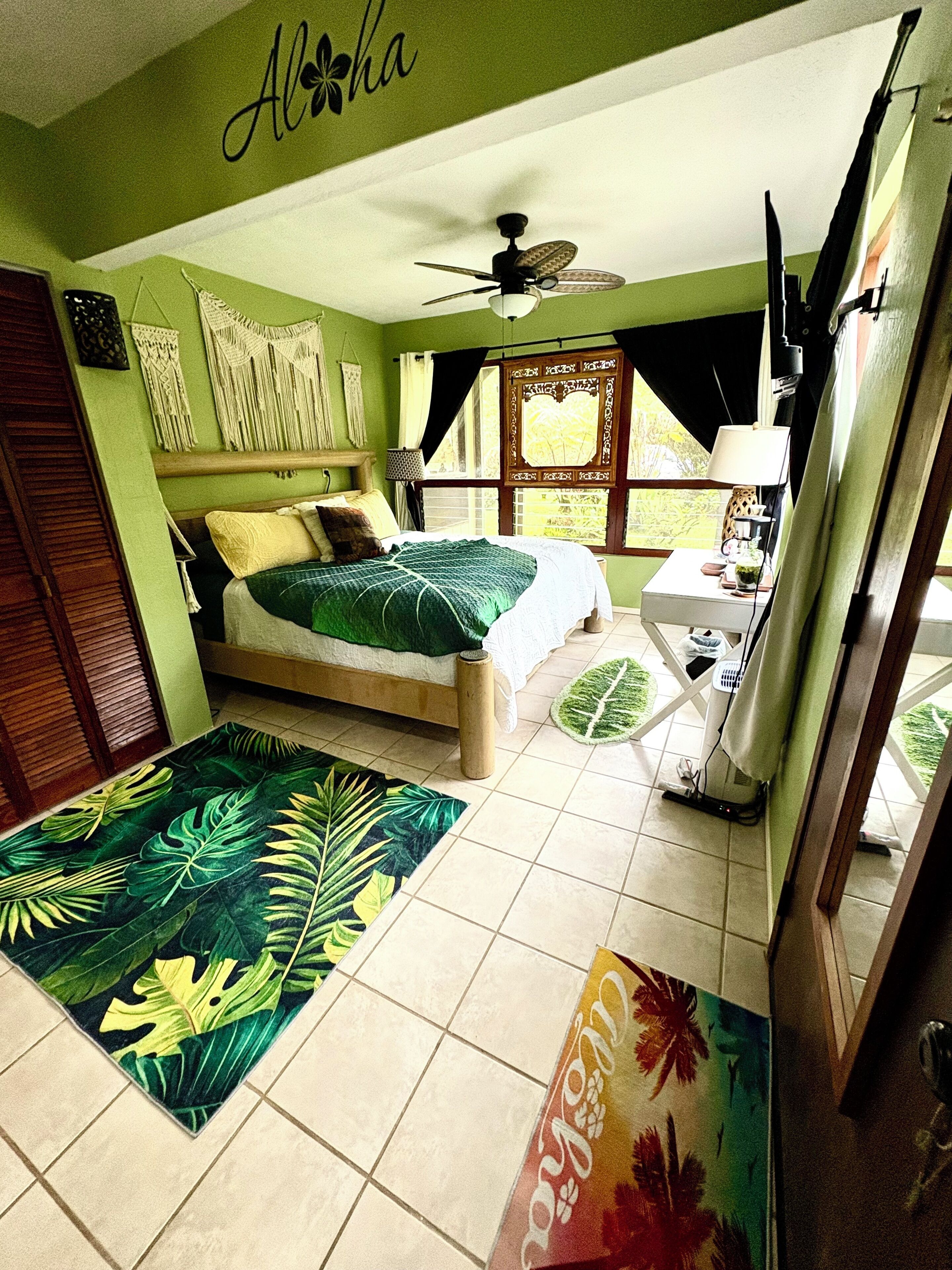 A bedroom with a bed, a ceiling fan, a window with curtains, a bedside table with a lamp, and a tropical-themed rug.
