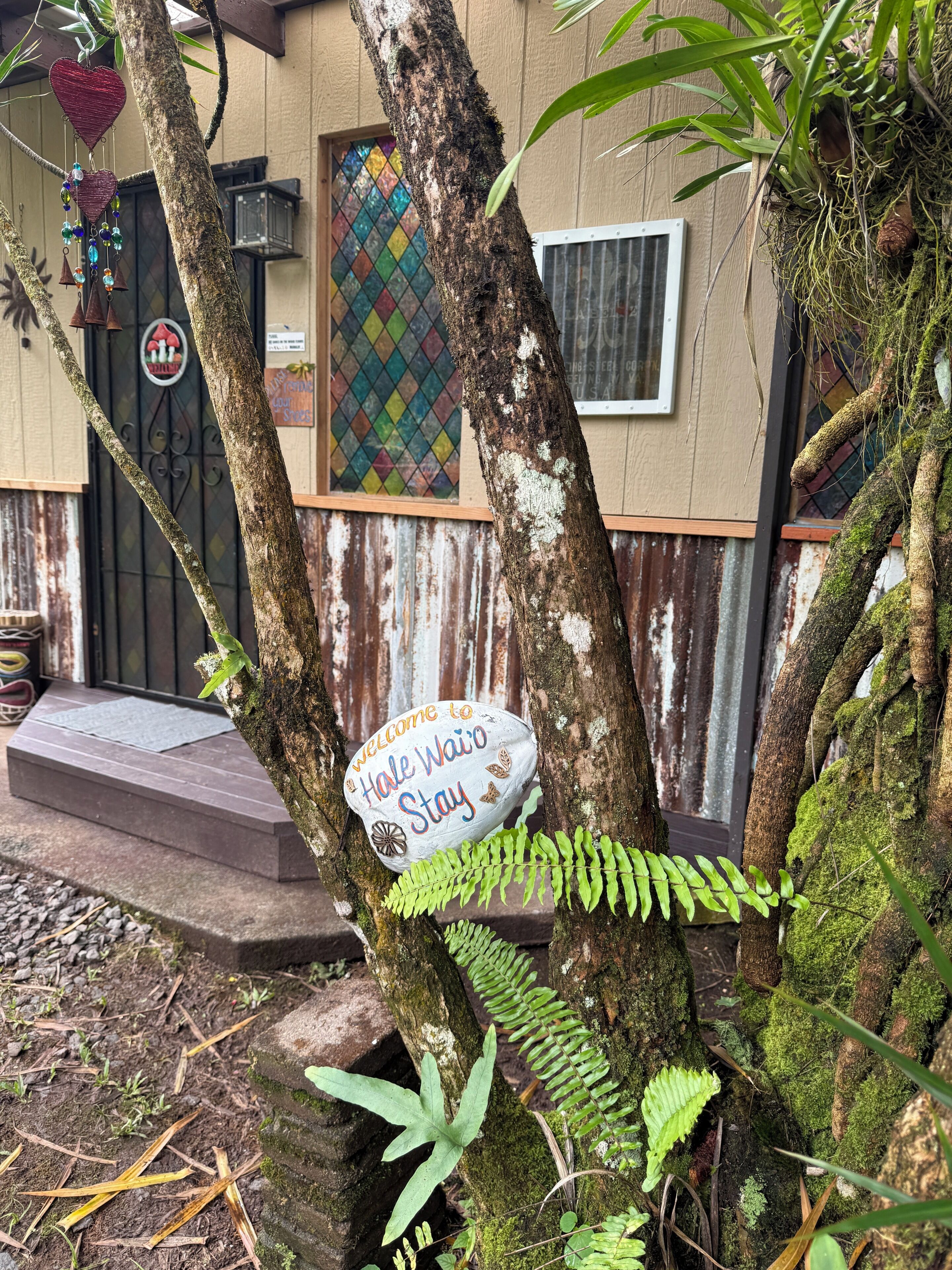 A tree with a 'Welcome to Hale Wai'o Stay' sign, a colorful stained glass window, and a rustic building facade.