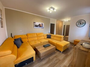 A modern living room with a yellow sectional sofa, a wooden coffee table, and a clock on the wall. - Vacation Rental No. 5, 50 square meters, 1 bedroom, bay window, terrace (Oberaudorf)