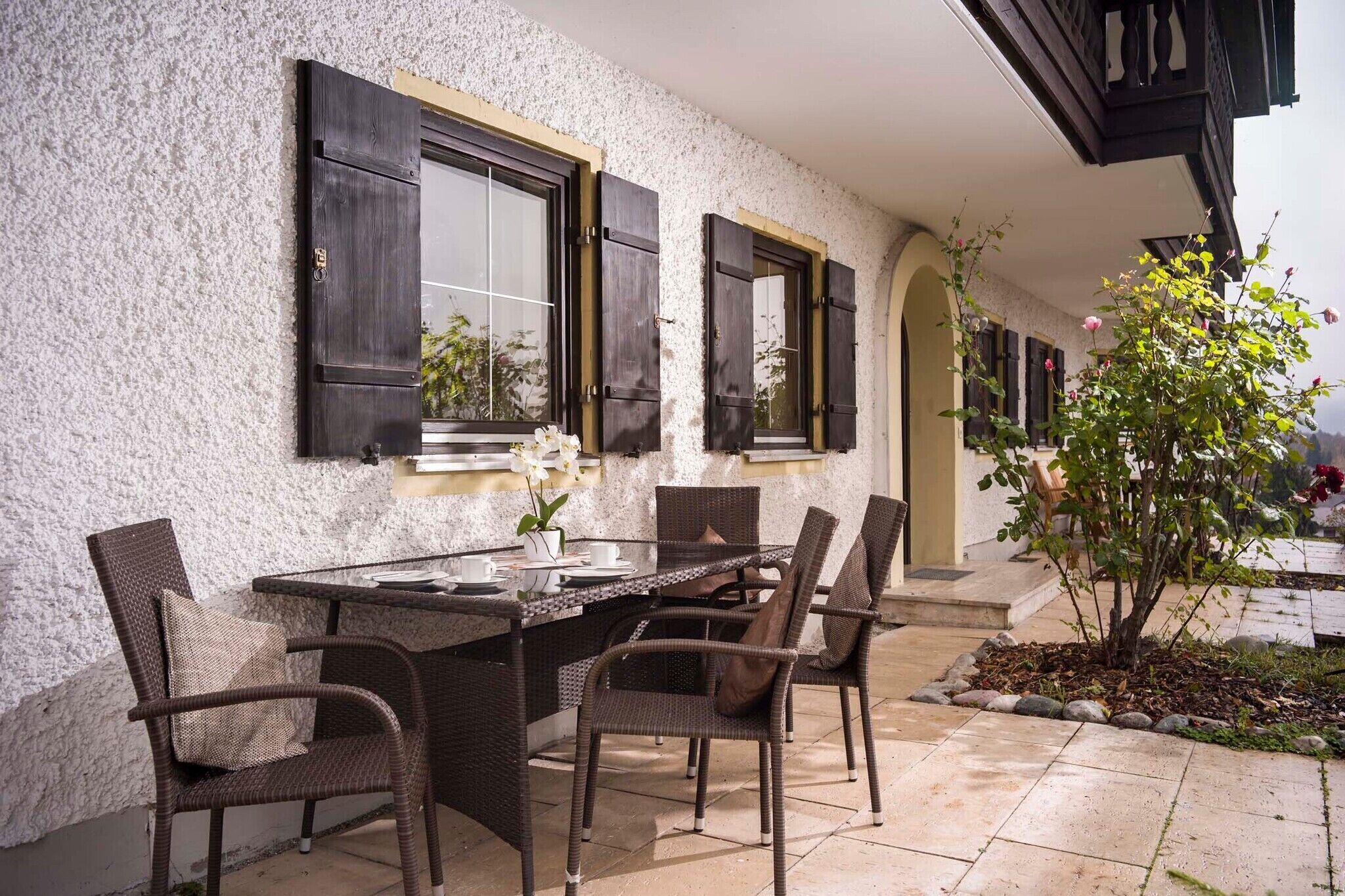 An outdoor dining area with a table and chairs, a window with shutters, and a small garden with a tree and flowers.