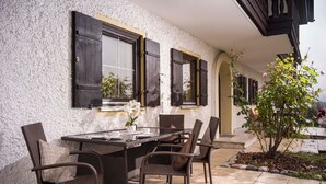 Ein Außenbereich zum Essen mit Tisch und Stühlen, ein Fenster mit Rollläden und ein kleiner Garten mit Baum und Blumen.