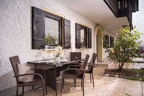 Ein Außenbereich zum Essen mit Tisch und Stühlen, ein Fenster mit Rollläden und ein kleiner Garten mit Baum und Blumen.