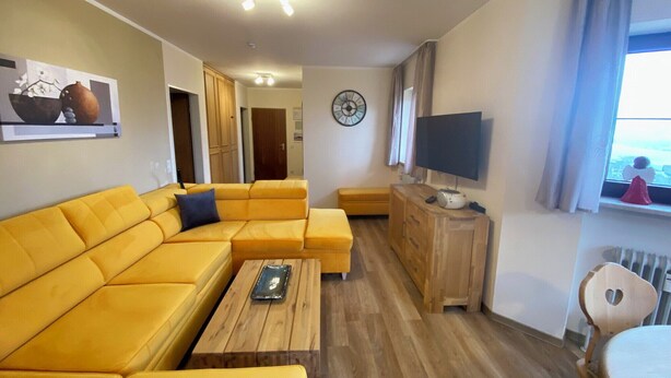 Ein Wohnzimmer mit gelber Couch, einem Holztisch, einem Fernseher auf einem Holzschrank und einer Uhr an der Wand.
