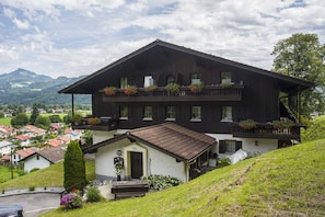 A two-story chalet with a balcony, a small attached house, and a view of a valley and mountains.