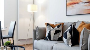 Un soggiorno moderno con un divano grigio, cuscini decorativi, una lampada da terra e un tavolino da caffè in vetro con oggetti ornamentali.