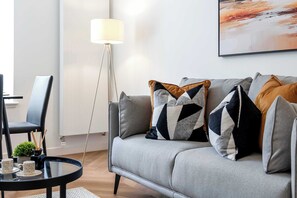 Ein stilvolles Wohnzimmer mit grauem Sofa, gemusterten Kissen, einer Stehlampe und einem Glastisch mit Dekoration.