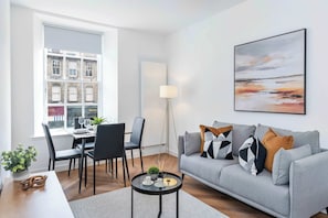 A modern living room with a grey sofa, a dining table with chairs, a coffee table, and a large window with a view of buildings. - Stylish city-centre two-bedroom apartment (Edinburgh)