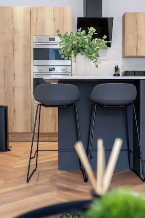 A modern kitchen with wooden cabinets, a built-in oven, a black countertop, and two black bar stools. - Stylish city-centre two-bedroom apartment (Edinburgh)