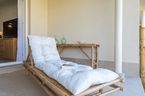 Ein Holz-Loungesessel mit weißem Kissen, ein kleiner Tisch mit Flaschen und einer Schüssel, und eine Küche im Hintergrund.