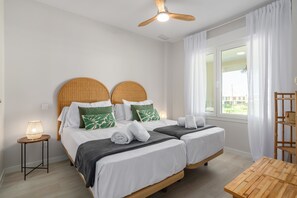 Ein Hotelzimmer mit zwei Betten, einem Deckenventilator, einem Fenster mit Vorhängen, einem kleinen Tisch und einer Lampe.