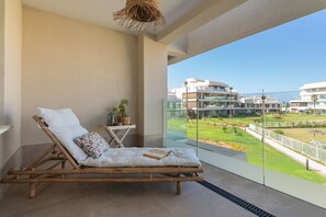 Een balkon met een rieten loungestoel, een klein tafeltje met een plant en vrij uitzicht op een wooncomplex.