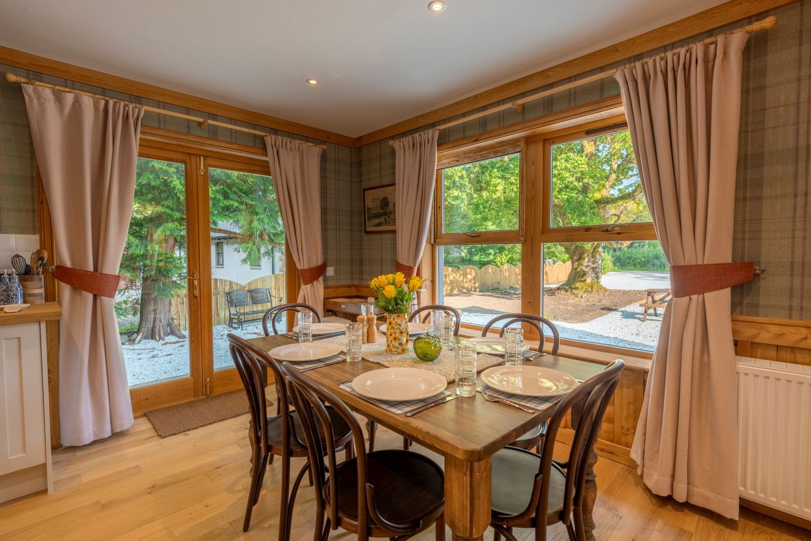Une salle à manger avec une table en bois dressée pour quatre personnes, de grandes fenêtres avec des rideaux et une vue sur l’extérieur.
