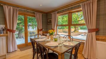 Une salle à manger avec une table en bois dressée pour quatre personnes, de grandes fenêtres avec des rideaux et une vue sur l’extérieur.