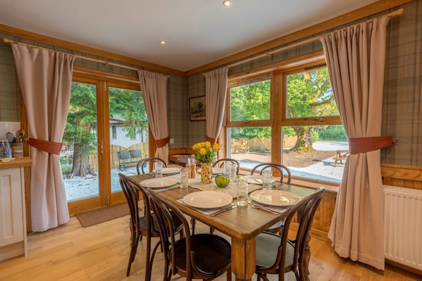A dining room with a wooden table set for four, large windows with curtains, and a view of the outdoors.