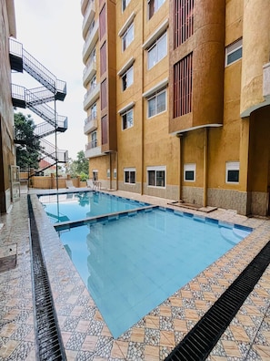 Una piscina con solárium empedrado, rodeada por un edificio de varios pisos con balcones y ventanas.