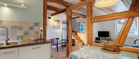 Un interior rústico con vigas de madera a la vista, una cocina con azulejos estampados, un comedor con mantel floral y una escalera que conduce a otro nivel.