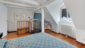 Un dormitorio con una cama, un cofre de madera, una ventana con persianas y una silla con cojín.