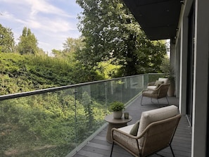 A balcony with wicker furniture, a small table with a plant, and a glass railing overlooking a lush green landscape. - 1 & 2 Bed Chic Apartments near Battersea Central London FREE WIFI & AIRCON (London)