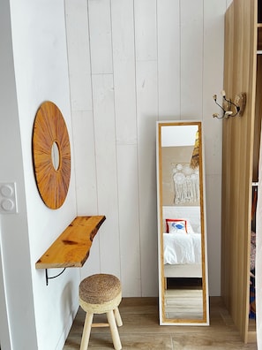 Een kamer met een houten vloer, een witte muur, een ronde spiegel, een houten plankje, een krukje en een grote spiegel die een bed met een witte dekbedovertrek en decoratieve kussens weerkaatst.