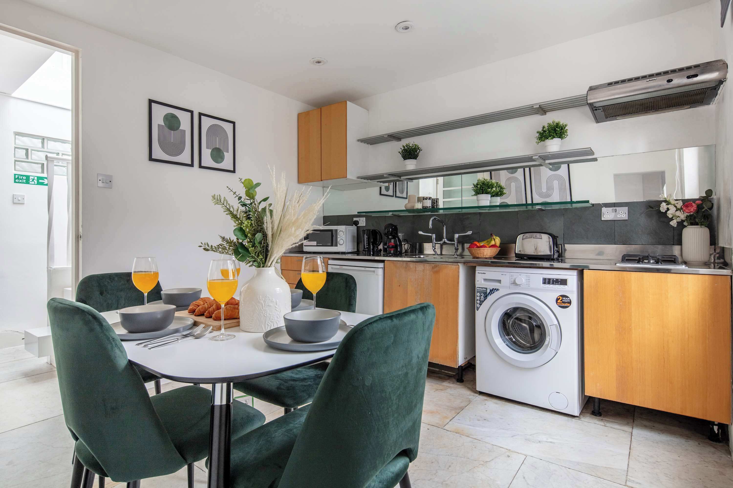 A modern kitchen with a washing machine, a dining area with green chairs, and a table set with glasses and a bowl.