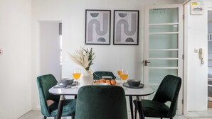Uma área de refeições com uma mesa redonda, cadeiras verdes e um vaso com plantas secas. Há duas obras de arte abstratas emolduradas na parede, uma porta com painéis de vidro fosco e um quadro branco com sinal de alerta.