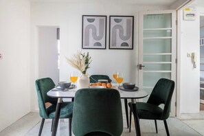Ein Essbereich mit einem runden Tisch, grünen Stühlen und einer Vase mit getrockneten Pflanzen. An der Wand hängen zwei gerahmte abstrakte Kunstwerke, eine Tür mit satinierten Glasfeldern und eine Tafel mit einem Warnhinweis.