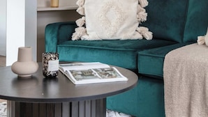 Um sofá azul-esverdeado com um cobertor branco, uma mesa redonda com um vaso e um livro, e uma estante branca com objetos decorativos.