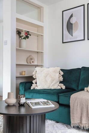 Ein türkissovas Sofa mit weißer Decke, ein runder Tisch mit einer Vase und einem Buch sowie ein weißes Regal mit Dekorationselementen.