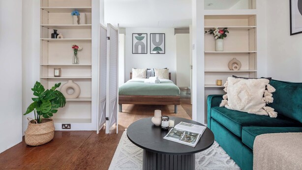 Uma sala de estar moderna com um sofá verde, uma mesa de centro redonda e uma cama com cabeceira verde. Há prateleiras com objetos decorativos e um vaso de planta.