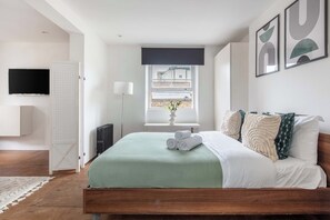Een slaapkamer met een bed, een raam, een radiator, een televisie en kunst aan de muur.